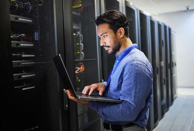 A man in blue shirt using laptop near server racks.